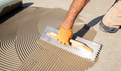 Construction worker spreading tile adhesive on floor using notched trowel, preparing surface for tiling