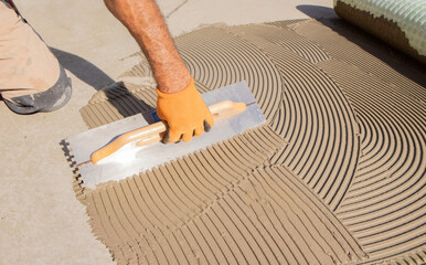 Construction worker spreading tile adhesive on floor using notched trowel, preparing surface for...