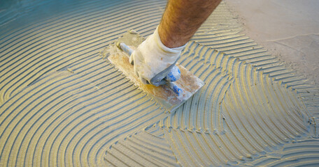 Construction worker spreading tile adhesive on floor using notched trowel, preparing surface for...