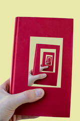A hand is a holding red book which in it's cover has a photo of the same hand holding the same book.