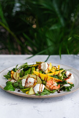 A tropical salad featuring fresh mango slices, shrimp, creamy burrata, arugula, and avocado.