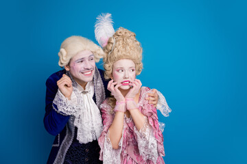 Stylish couple in baroque costume posing humorously against a blue background, showcasing historical elegance