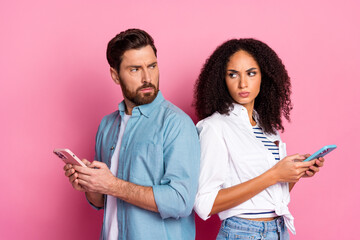 Young couple in a casual style with smartphones looking suspiciously at each other, set against a vibrant pink background