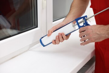 Worker with caulking gun sealing window indoors, closeup