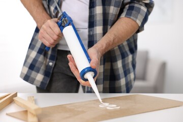 Man with caulking gun glueing plywood at white table indoors, closeup