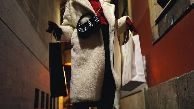 Happy curly girl runs from shopping with gift bags in her hands, impressionable, consumption, shops, shopping, dance, energy, movement, money, holidays, night, city alley, fashion, clothes, details