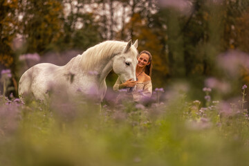Fototapeta premium A young woman cuddle and interact with her white spanish horese on a wildflower field in autumn outdoors