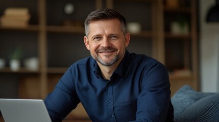 Fototapeta premium Man with short beard smiles while sitting at his desk with a laptop in a contemporary workspace