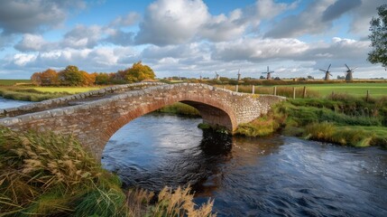 Obraz premium Stone arch bridge over calm river, autumnal landscape with windmills.