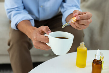 Young man dripping CBD tincture into drink at white table, closeup