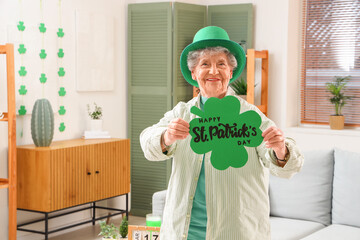 Senior woman in leprechaun hat holding festive postcard with text HAPPY ST. PATRICK'S DAY at home