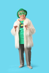 Happy senior woman in fur coat, glasses and leprechaun hat for St. Patrick's Day celebration on blue background