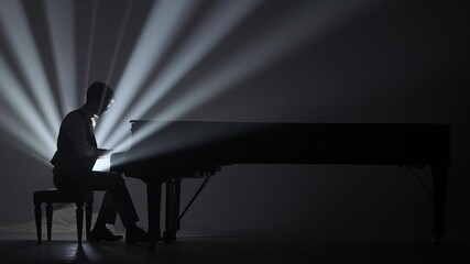 Pianist sitting and playing on the black grand piano on dark concert stage against bright spotlight, isolated on black studio background. Artist performing in concert hall.