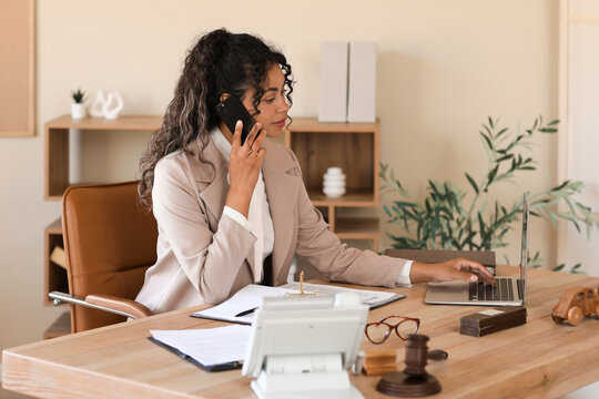 Female African-American lawyer with laptop talking by mobile phone at table in office
