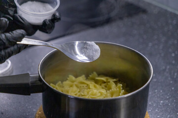 Adding salt to farfalle pasta in a metal pot using a spoon on a kitchen countertop