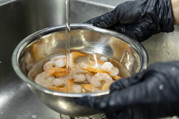 fresh shrimp being rinsed in stainless steel bowl under running water by hands in black gloves in...