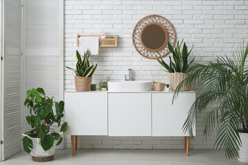 Modern sink, chest of drawers, houseplants and folding screen near white brick wall in bathroom