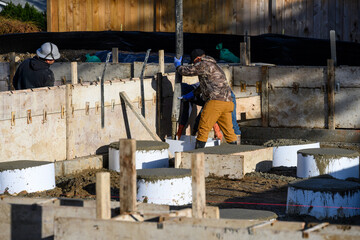 Workmen filling cement into round and square plastic forms for house foundation footings with a pipe from a Concrete Boom Pump Truck, worker with trowel smoothing out cement
