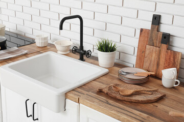 Fototapeta premium White sink, chopping board and kitchen utensils on counter near white brick wall. Closeup