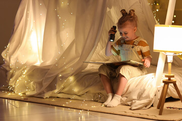 Cute little girl with flashlight reading story in hut at home