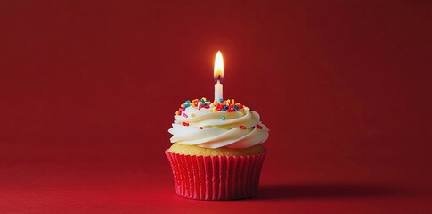 A cupcake with white frosting and colorful sprinkles, topped with one lit birthday candle on a red background