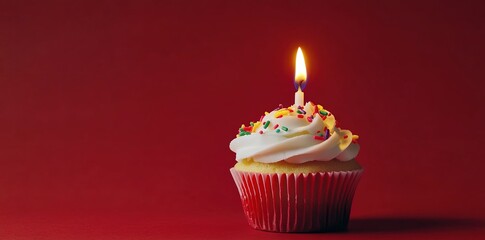 A cupcake with white frosting and colorful sprinkles, topped with one lit birthday candle on a red background