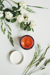 Close up of face cream in open glass jar and blooming flowers on white background. Set of natural cosmetics for skin or body care beauty products