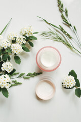 Close up of face cream in open glass jar and blooming flowers on white background. Set of natural cosmetics for skin or body care beauty products