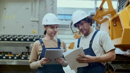Female technician in hard hat and uniform, standing in front of heavy machinery, using digital tablet and discussing paper plans with male coworker