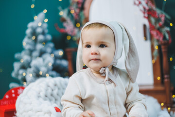 A 1 year old boy sits near a Christmas tree in a bunny costume. New Year's celebration for children