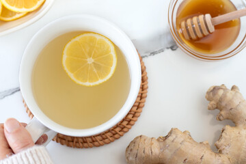 Hand holding cup of ginger lemon tea on white marble table with honey. Top view. Warming and relaxing with homemade hot drink.