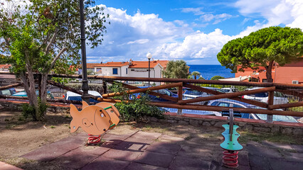 The element of the playground with swings on the spring, rocking chair. In the background - houses, sea and blue sky with white clouds. Pomonte, Elba, Tuscany, Italy.
