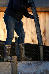 Construction worker walking on wood cement form panels for foundation walls guiding concrete boom pump truck pipe into forms, winter house building project job site
