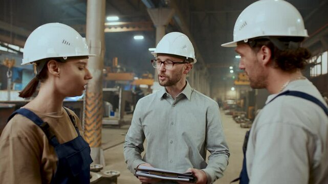 Engineer in hard hat holding documents and giving instructions to male and female technicians during workday in industrial plant full of heavy machinery