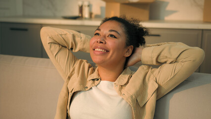 Happy relaxed African American woman female home owner joyful girl renter tenant sit on couch relax enjoy house renovation moving in new apartment relocation day buy rent real estate mortgage smile