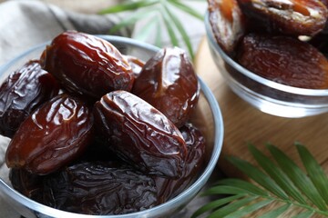 Many tasty dried dates in bowls and leaves on table, closeup