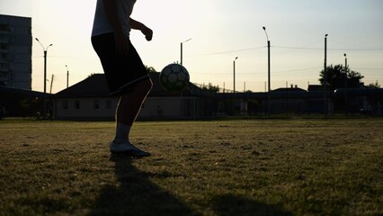 Legs of young man kicking ball at green field. Male feet of professional footballer juggling soccer ball on stadium at sunset. Sportsman practicing tricks outdoor. Concept of a freestyle football