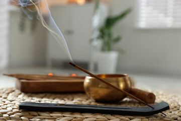 Incense stick smoldering in holder and tibetan singing bowl on wicker mat indoors