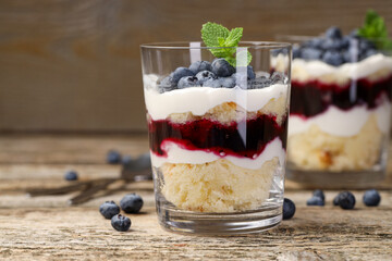 Tasty trifle dessert. Sponge cake, blueberries, jam and whipped cream in glasses on wooden table, closeup