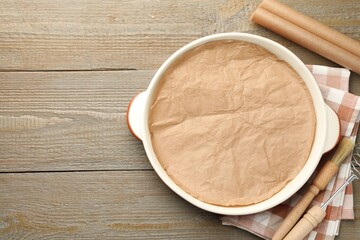 Ceramic casserole, baking parchment paper, brush and whisk on wooden table, flat lay. Space for text