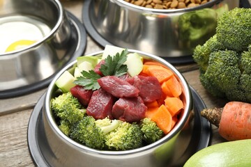 Fresh natural pet food in feeding bowl and vegetables on wooden table, closeup