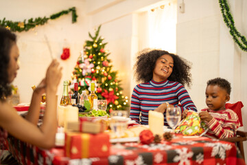A cheerful holiday dinner scene with a diverse family enjoying a festive meal. Christmas decorations, gifts, and a lit Christmas tree create a cozy atmosphere, enhancing the warmth of the celebration.