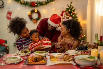 A joyful Christmas celebration with friends gathered around a festive dining table. Featuring a man in a Santa hat, a decorated Christmas tree, delicious food, and warm holiday cheer.