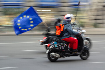 Fototapeta premium Unidentifiable man in red jacket in out of focus on motorcycle with EU flag with yellow stars. Street blurry defocused photo.Concept of transport, rally on city street, protest. Blurred background