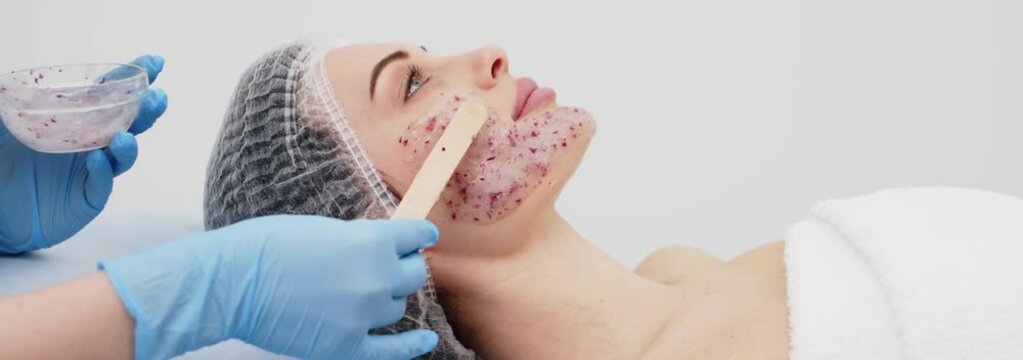 Close-up of a cosmetologist&rsquo;s hands applying an alginate mask with rose petals to a young woman&rsquo;s face in a professional beauty salon. Shot on anamorphic lens in 2.39