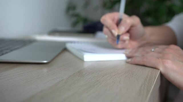 The woman is taking notes in a notebook and working on a laptop.