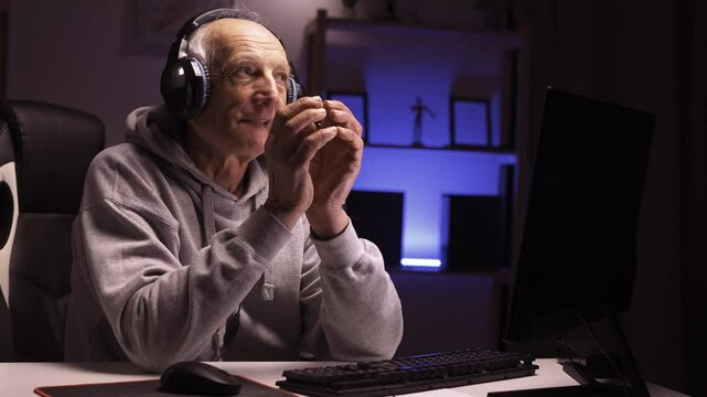 artful senior mature gamer wearing headphones streaming his nighttime gaming session using personal computer with fake sly and tricky expression, sitting at home gaming room.