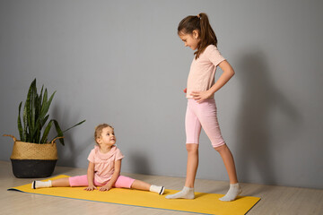 Two girls engage in yoga practice at home. One performs a split on a yellow mat, while the other...