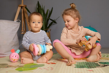 Two siblings engage with vibrant toys on a soft play mat in a warm indoor environment. The older child watches the younger one with interest, creating a joyful moment