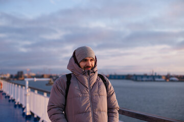 Mid section portrait of a handsome man wearing warm puffer coat standing on a sun deck of a cruise ship at sunny winter day against cloudy sunrise epic sky